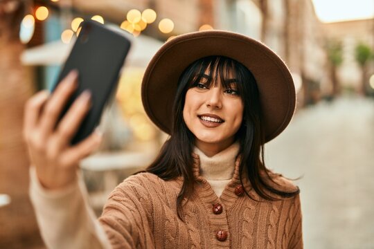 Brunette woman wearing winter hat taking a selfie picture with smartphone outdoors at the city