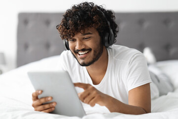 Cheerful hindu guy using digital tablet and headphones in bed