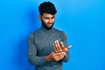 Arab man with beard wearing turtleneck sweater suffering pain on hands and fingers, arthritis...