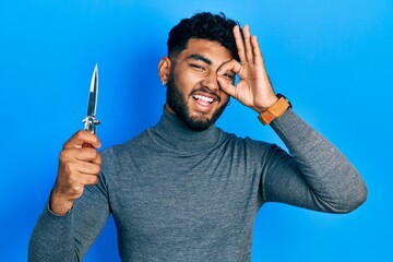 Arab man with beard holding pocket knife smiling happy doing ok sign with hand on eye looking through fingers