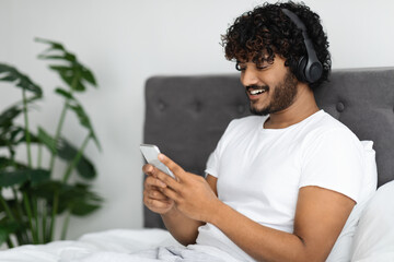 Happy indian guy sitting in bed with smartphone and headset