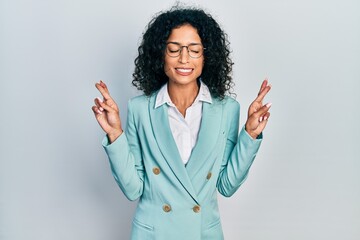 Young latin girl wearing business clothes and glasses gesturing finger crossed smiling with hope...