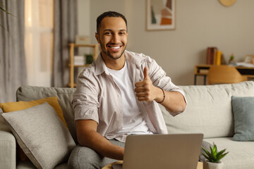 Happy young black guy working on laptop computer, showing thumb up, recommending remote job at home