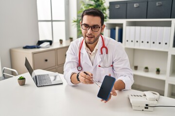 Fototapeta na wymiar Young hispanic man wearing doctor uniform showing medical smartphone app at clinic
