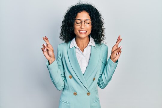 Young latin girl wearing business clothes and glasses gesturing finger crossed smiling with hope and eyes closed. luck and superstitious concept.
