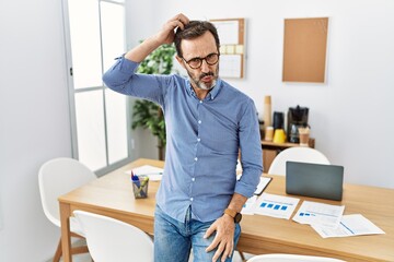 Middle age hispanic man with beard wearing business clothes at the office confuse and wonder about question. uncertain with doubt, thinking with hand on head. pensive concept.