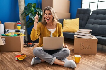 Young woman sitting on the floor at new home using laptop pointing finger up with successful idea. exited and happy. number one.