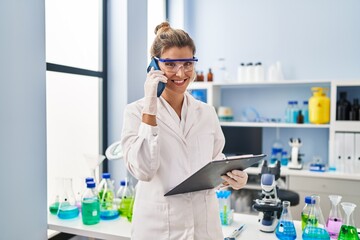 Young blonde woman wearing scientist uniform talking on the smartphone at laboratory