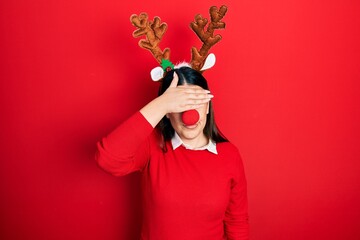 Young hispanic woman wearing deer christmas hat and red nose covering eyes with hand, looking serious and sad. sightless, hiding and rejection concept