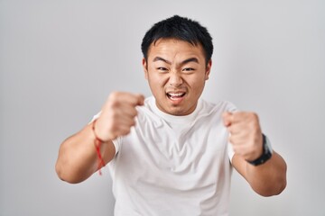 Young chinese man standing over white background angry and mad raising fists frustrated and furious while shouting with anger. rage and aggressive concept.