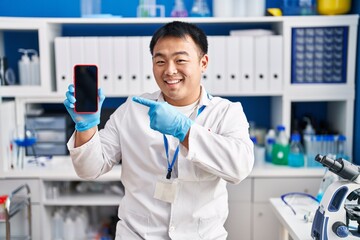 Young chinese man working at scientist laboratory holding smartphone smiling happy pointing with hand and finger