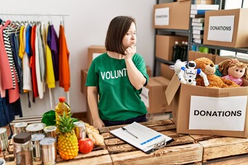 Young down syndrome woman wearing volunteer t shirt at donations stand serious face thinking about...