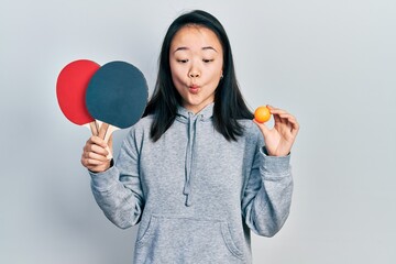 Young chinese girl holding red ping pong rackets and ball making fish face with mouth and squinting...