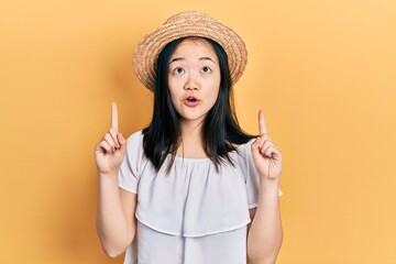 Young chinese girl wearing summer hat amazed and surprised looking up and pointing with fingers and raised arms.