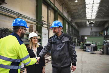 Engineer and industrial worker in uniform shaking hands in large metal factory hall and talking.