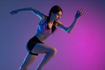 Fototapeta na wymiar One young muscular girl, female runner or jogger training isolated on pink-blue background in neon light. Sport, track-and-field athletics, competition and active lifestyle concept