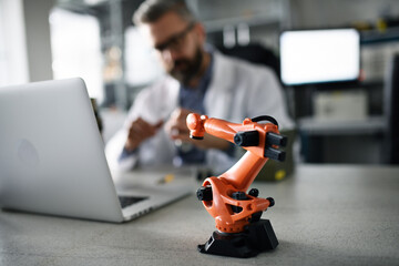 Robot arm industrial miniature figure on table in front of robotics engineer working on laptop in...