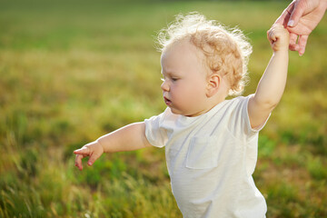 First baby steps. A young mother helps a baby boy walk on the grass in the park. Parental care and support