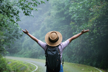 Happy female traveler in a hat with a backpack walks on the road in the rainforest. Female traveler...
