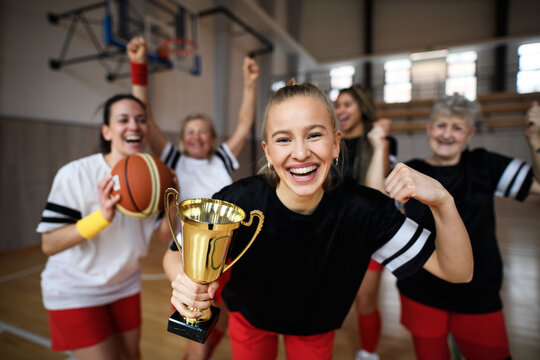 Group Of Young And Old Women, Basketball Team Players, In Gym With Trophy Celebrating Victory.