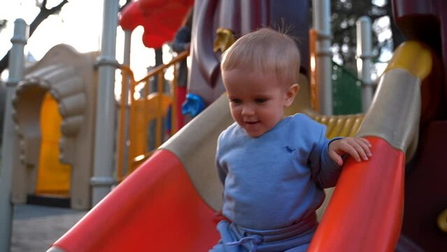 Little kid boy slides down colorful slide installed at playground in urban park on sunny day. Cute toddler enjoys spring activity closeup slow motion