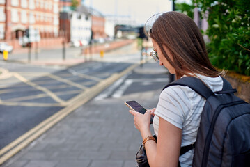 Fototapeta premium The girl looks at the phone standing on the sidewalk. Writes sms. A girl with a backpack looks at the navigator, standing on the street.