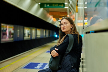 A girl with glasses is waiting for a train in the subway, sitting on a bench and hugging her backpack.