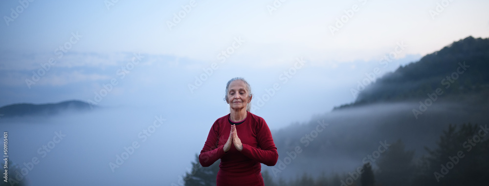 Wall mural Senior woman doing breathing exercise in nature on early morning with fog and mountains in background.