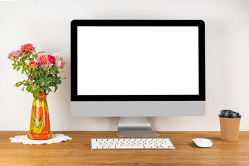 Blank screen desktop computer with wireless keyboard and mouse on wooden table in a working room with a vase of roses flower and a peper cup of coffee.