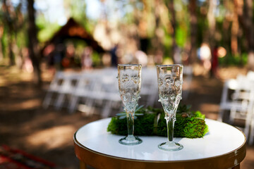 Wine glasses, wedding rings bedding of grass and moss on white table next to bohemian tipi arch...