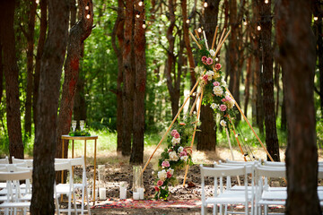 Beautiful bohemian tipi arch decoration on outdoor wedding ceremony venue in pine forest with cones. Chairs, floristic flower compositions of roses, carpet, string fairy lights. Summer rural wedding. © artiemedvedev