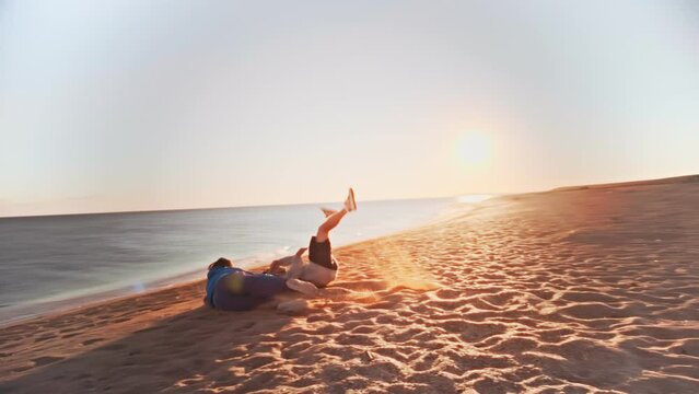Young guy starts dancing trendy dance on the beach by the sea against the background of setting sun. The girl comes running and knocks the guy off his feet and both fall on the sand.