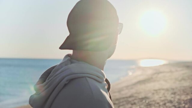 Rear view of young man in sunglasses and baseball cap looks at the sunset standing on sandy beach by the sea. Happy guy enjoys the beautiful seascape.