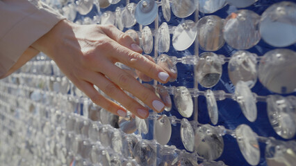 Female hand with white manicure touches city wall decorated with sequins. Woman walks along sparkling facade during city stroll and touches sequins