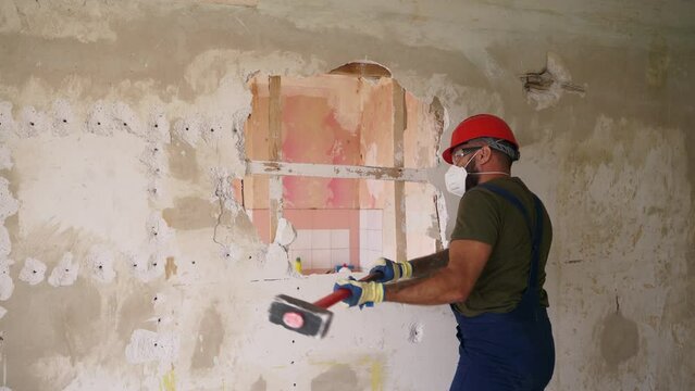 Contractor wrecks wall with sledgehammer making hole for rearrangement. Man doing manual dismantling and demolition works with big hammer hits for apartment renovation. Construction worker in uniform.