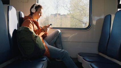 Young man travels alone by electric train with backpack. Man in white wireless headphones scrolls...