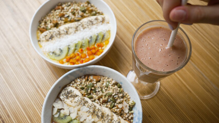 Sweet vegan cereal bowls and fruit smoothie stand on wooden table for breakfast. Female hand stirs fresh fruit smoothie with white straw in cafe closeup