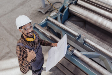 High angle view of heavy industry worker with safety headphones and hard hat in industrial factory holding blueprints