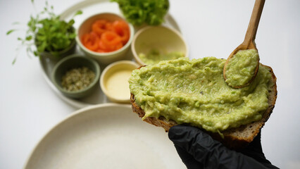 Cook in black rubber gloves spreads guacamole on toast with wooden tablespoon. Ingredients stand on background. Process of cooking closeup
