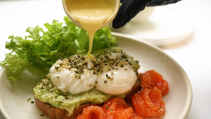 Chef pours sauce on toast with guacamole and poached eggs sprinkled with seeds. Trout and fresh salad leaves served on side of toast closeup