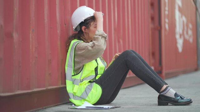Asian cargo container worker woman sit on floor and lean to the tank with relaxing also use clipboard to mank wind for releasing hot condition in workplace.