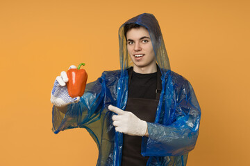 Young male gardener in a blue raincoat and gloves holds a red pepper in his hand on a yellow background