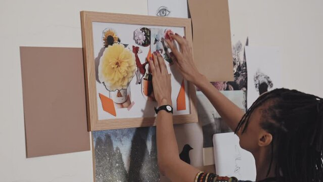Close up view of African American woman attaching paper element to canvas in flame while making creative collage on wall in art studio