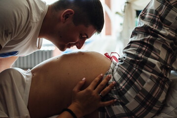 Image of happy future dad kissing belly of his cheerful caucasian pregnant wife lying on bed at home, close up. Film noise