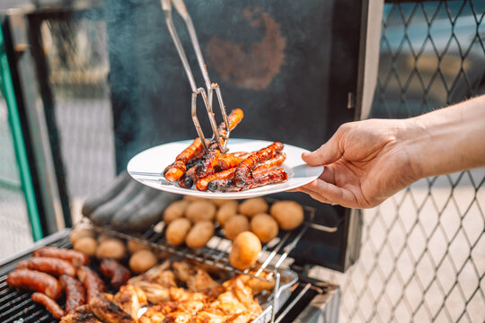 Closeup View Of A Hot Food Vendor Cooking Pork Sausages And Chops Over A Hot Grill With Rising Steam, Chef Uses Tongs To Flip Meat At Local Agricultural Fair