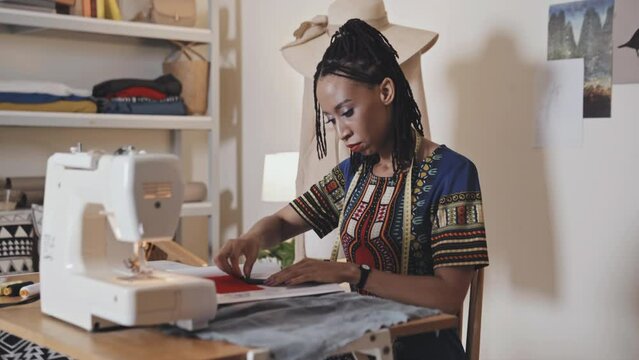 Young African American woman sitting at desk with sewing machine and working with fabric in tailor studio