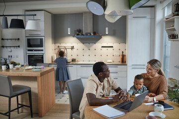Wide angle portrait of black father working from home with wife and child sitting at table beside him in cozy home scene, copy space