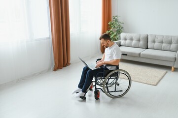 Portrait of smiling disabled male sitting in wheelchair and working on laptop from home. Young worker with special needs. Freelancer and people with disabilities concept