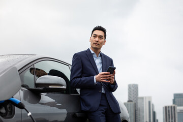 Businessman using smartphone and charging electric car in the city