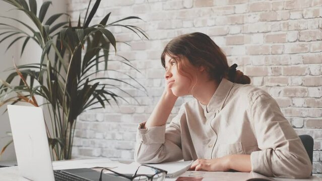 Young woman student sitting at desk full with books textbooks looking at computer screen to unfinished homework feels tired bored and unmotivated, girl loaded with tasks, work backlog overflow concept